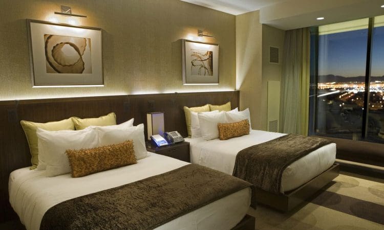 Modern hotel room with two double beds, brown and gold bedding, abstract art above the beds, large window with a city night view, and neutral decor.
