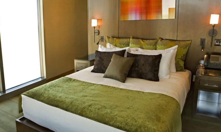 Modern hotel room with a neatly made bed, green and brown pillows and throw, wooden headboard, bedside tables, wall lamps, phone, and a colorful abstract painting above the bed.