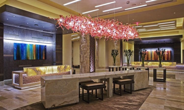 Modern hotel lobby featuring a long marble table, cushioned stools, abstract art, decorative trees, and a large overhead light fixture with pink glass elements.
