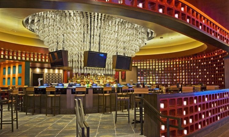 A modern bar with a large, sculptural chandelier, multiple hanging TVs, bar stools, and a circular counter surrounded by geometric wall designs.