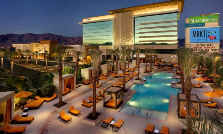 Outdoor view of Aliante Casino and Hotel at dusk, featuring a modern building, illuminated pool area, orange lounge chairs, palm trees, and mountains in the background.