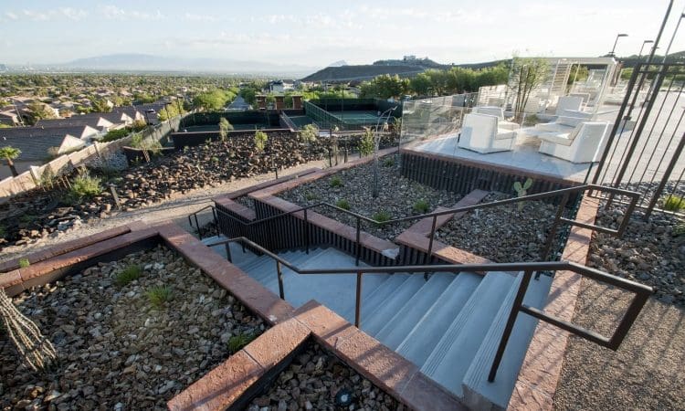 A concrete staircase with metal railings leads down through a landscaped area with rocks and desert plants, overlooking a residential neighborhood and distant mountains.