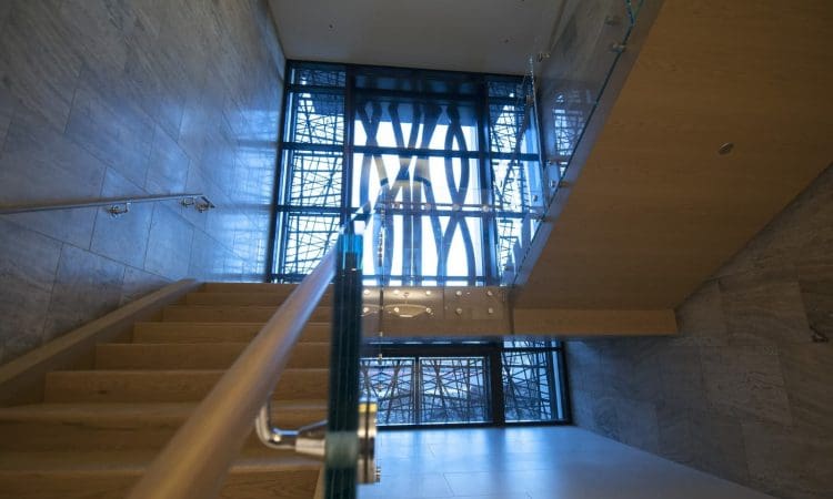 Modern indoor staircase with wooden steps and glass railing, leading up to a large window with geometric metal patterns and a view of an exterior structure.