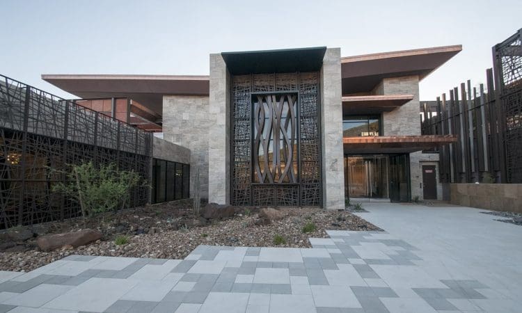 Modern building with a geometric facade, metal and stone accents, large decorative metal door, and a landscaped courtyard with rocks and sparse plants.