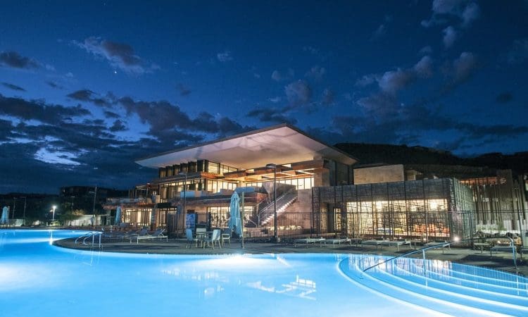 Modern building with large glass windows and outdoor lighting beside a curved swimming pool at dusk, under a partly cloudy sky.
