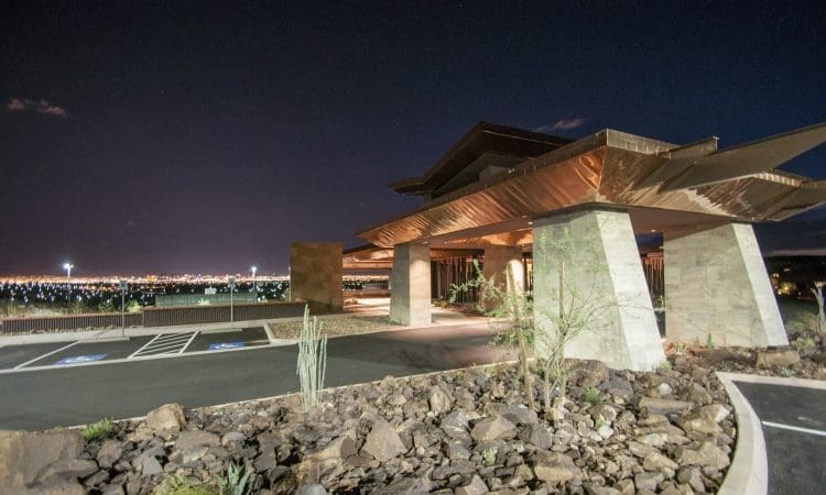 A modern building with angled copper roof and stone pillars is illuminated at night, with city lights visible in the distance and desert landscaping in the foreground.