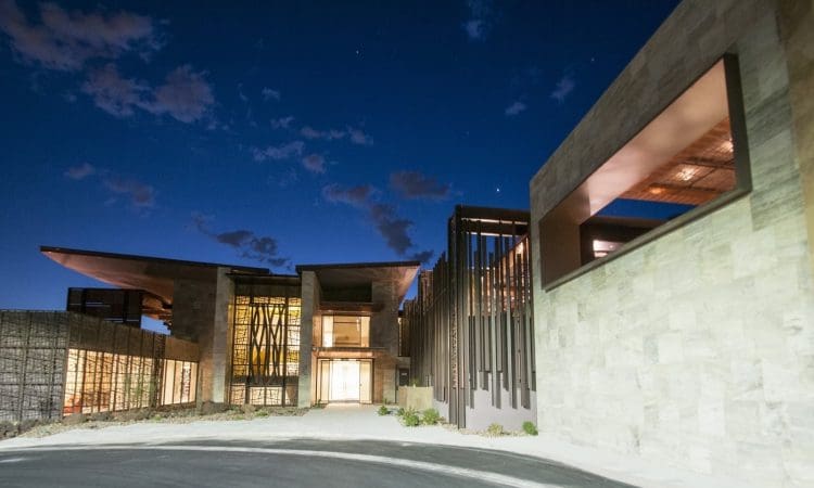 Modern building with large windows and angular rooflines, illuminated at night under a dark blue sky with scattered clouds.