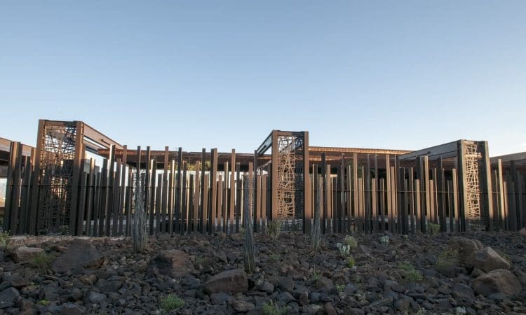 A modern building with vertical metal slats and decorative panels stands behind a rocky landscape with cacti under a clear sky.