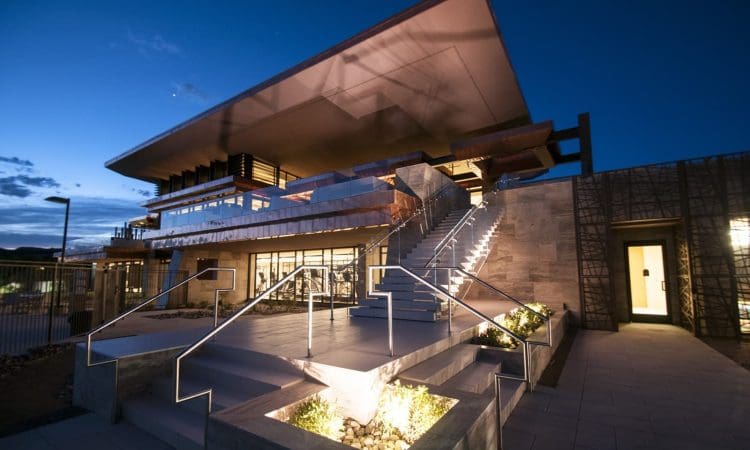 Modern building with large overhanging roof, illuminated steps, and glass walls, photographed at dusk with lights highlighting architectural features.