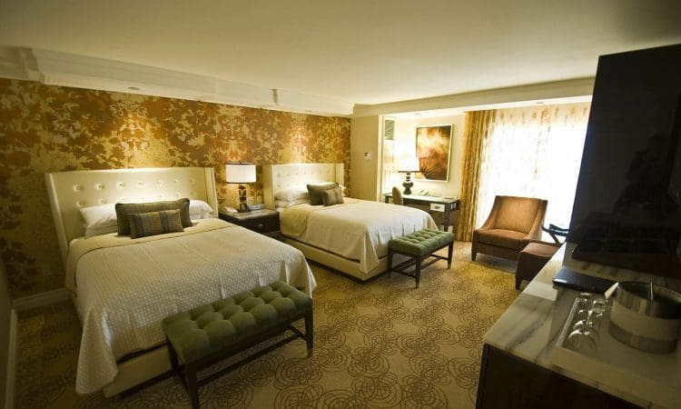 A hotel room with two double beds, green benches at each bed, patterned carpet, gold floral wallpaper, and a seating area by a large window with sheer curtains.