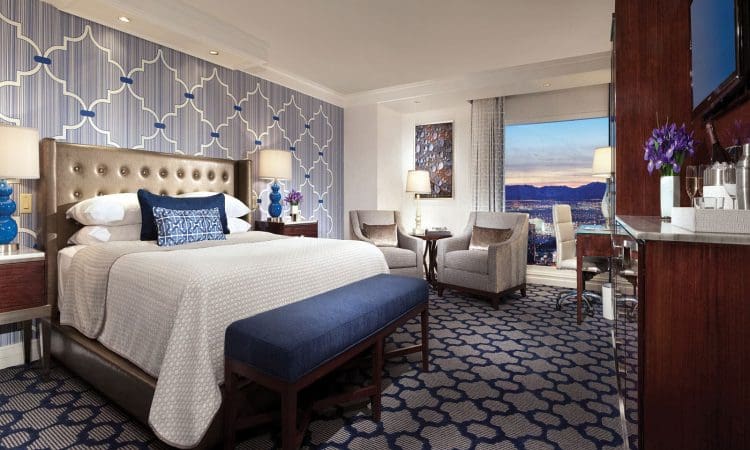 Modern hotel room with a large bed, blue bench, patterned wallpaper and carpet, seating area by a window, and a view of the city skyline at sunset.