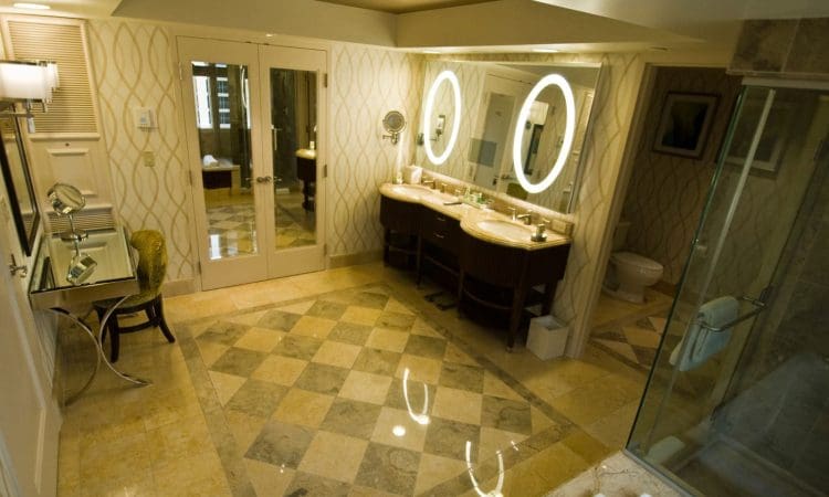Spacious bathroom with tiled floor, double sink vanity with illuminated mirrors, glass shower, toilet, and dressing area with a chair and wall mirror.