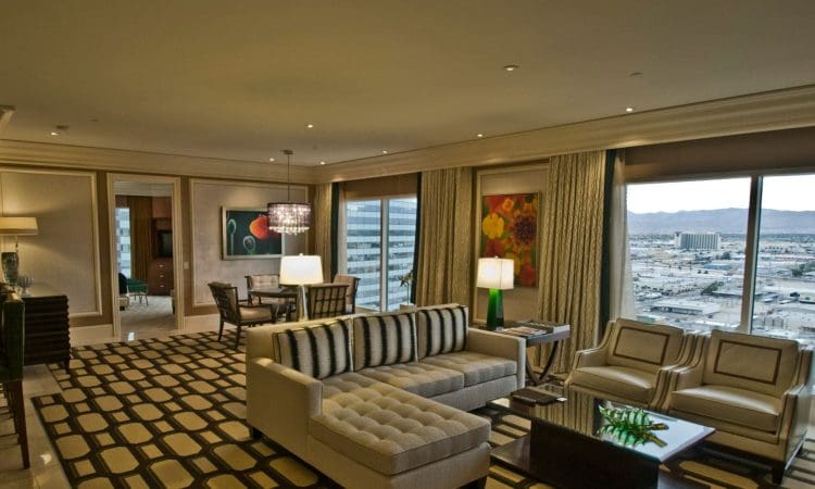 A modern hotel suite features a sectional sofa, two armchairs, patterned carpet, dining table, large windows with city and mountain views, and contemporary artwork on the walls.