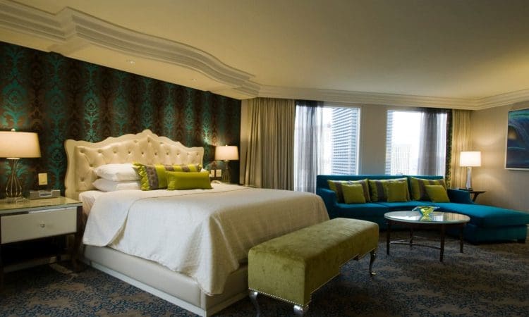 A hotel room with a large bed, decorative pillows, two nightstands with lamps, a bench, a blue sofa, a glass coffee table, and windows with city views.