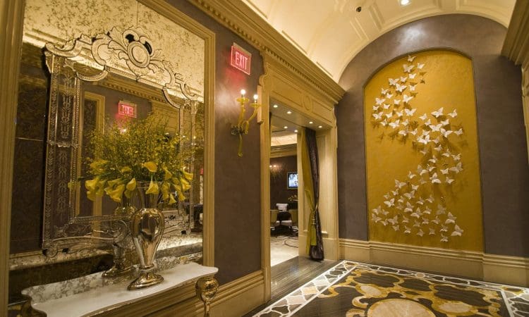 Elegant hallway with ornate mirror, marble-topped table holding a vase of yellow flowers, gold wall sconce, patterned rug, and decorative wall with white butterflies.