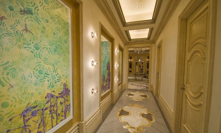 A hallway with beige walls, abstract green artwork, wall lights, patterned flooring, and a decorative ceiling, leading to a room with a chandelier.