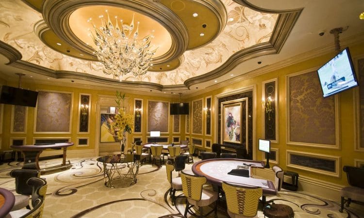 Luxurious casino room with ornate ceiling, chandelier, several gaming tables, chairs, wall art, and flat-screen TVs. Room is empty of people.