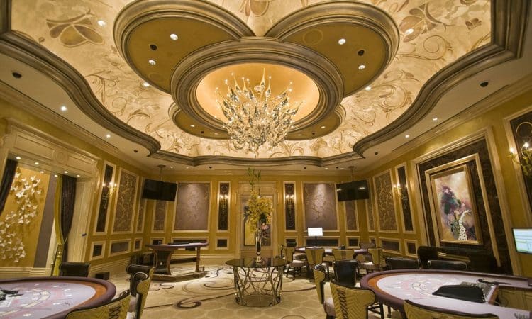 Elegant casino gaming room with ornate ceiling, chandelier, round tables, decorative wall art, and plush chairs arranged around the room.