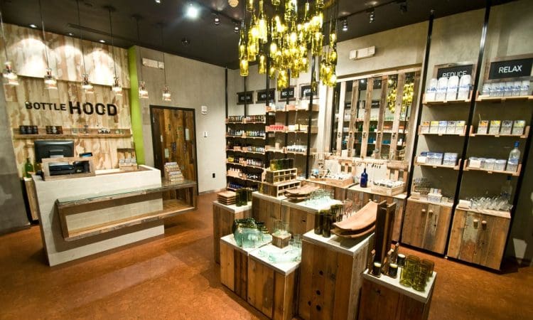 A modern store interior featuring wooden shelves and displays filled with glassware, bottles, and eco-friendly products; a chandelier made of bottles hangs from the ceiling.