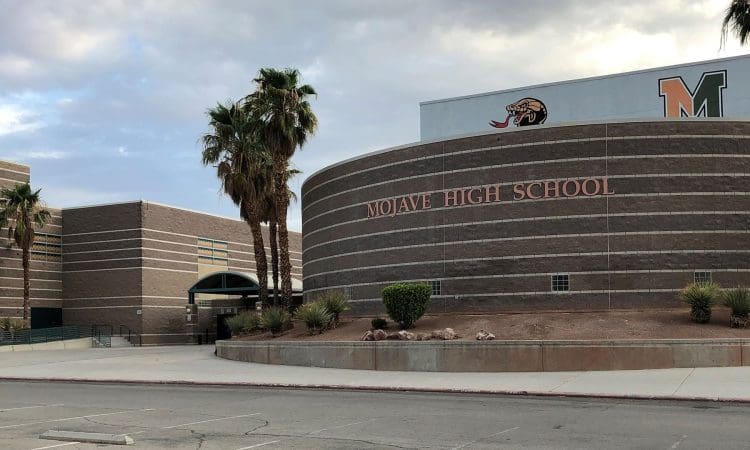 Curved brick building with "Mojave High School" sign, palm trees, and school mascots on the wall, under a cloudy sky.