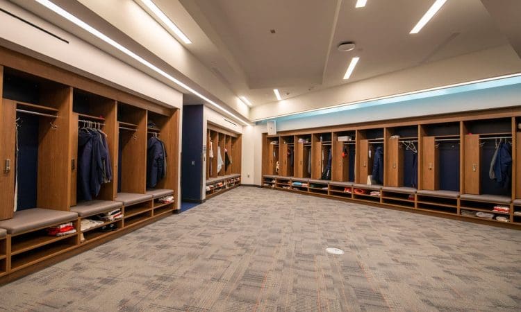 Spacious locker room with open wooden lockers, benches, and clothes hanging neatly; bright overhead lighting and carpeted floor.