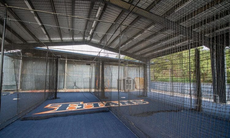 Indoor batting cage with netting, blue flooring, and a sports team logo painted on the ground under a metal roof structure.