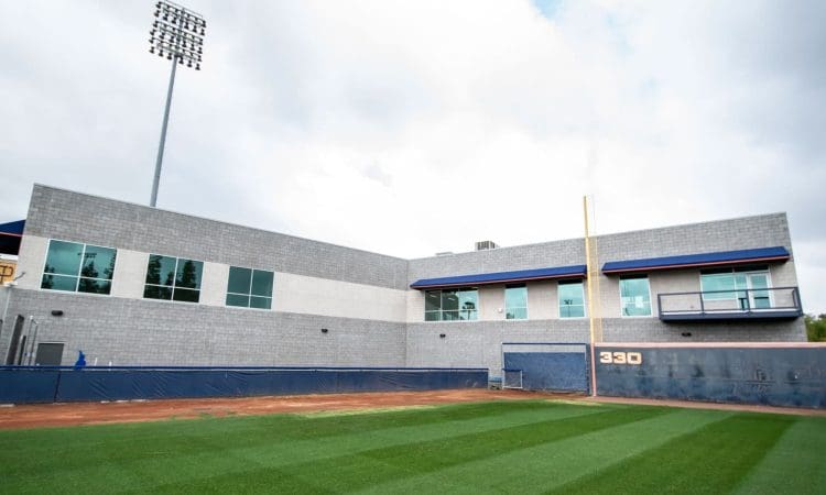 A view of a modern gray building with large windows behind the right outfield wall of a baseball field, featuring a 330-foot distance marker and a tall light pole.