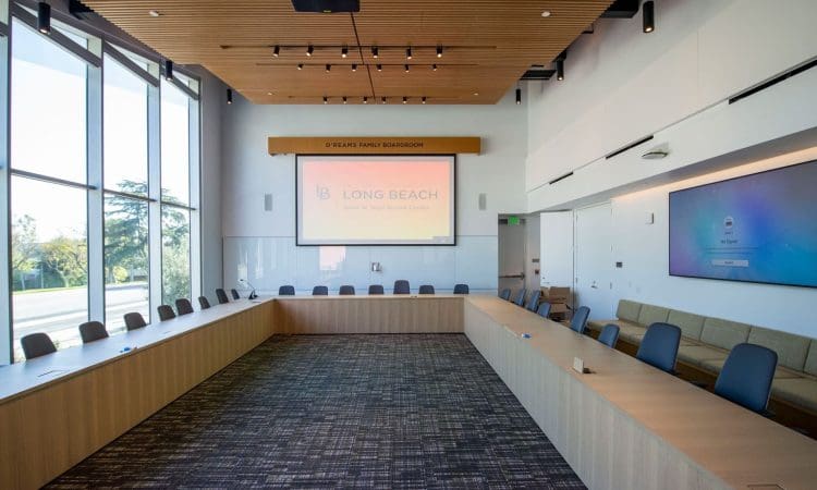 A modern conference room with a U-shaped table, chairs, large windows, wall-mounted screens, and a projector displaying "Long Beach" on the far wall.