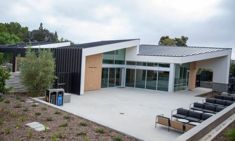 Modern building with large glass windows, angled roof, outdoor seating area, and landscaped surroundings under a cloudy sky.