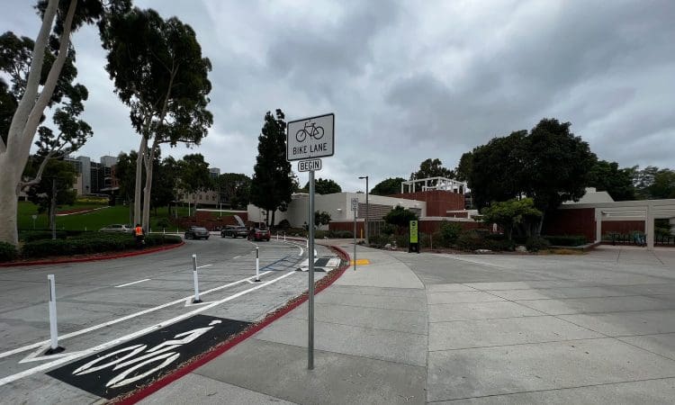 A bike lane with a sign reading "Bike Lane Begin" runs alongside a road bordered by trees, grass, and brick buildings under a cloudy sky.
