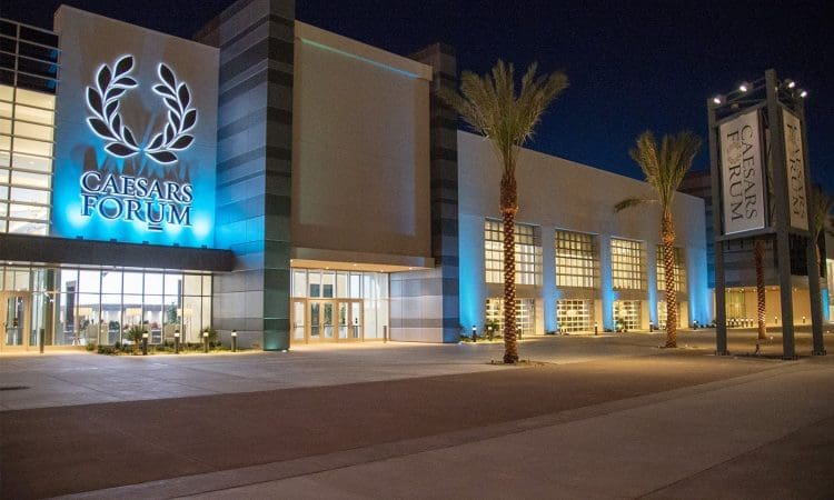 Exterior view of Caesars Forum convention center at night, illuminated with blue lighting and palm trees in front.