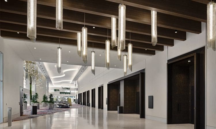 Spacious modern hotel hallway with glossy floors, dark wood ceiling beams, and multiple hanging cylindrical lights. Lounge area with seating and plants visible in the background.