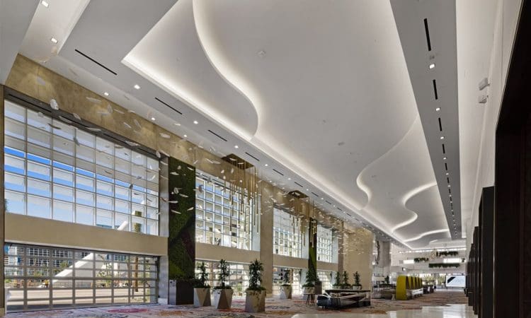 Spacious modern lobby with tall glass windows, wavy white ceiling, patterned carpet, plants, and seating areas, filled with natural light.