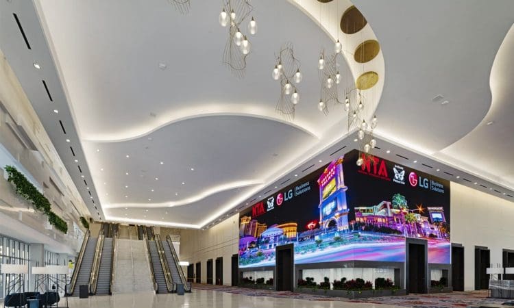 Spacious, modern convention center lobby with sweeping ceiling lights, escalators to the left, and a large digital display on the right wall.