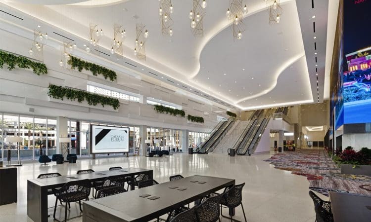 Modern lobby with high ceilings, decorative lighting, escalators, digital screen, long tables with chairs, and greenery accents on the walls.