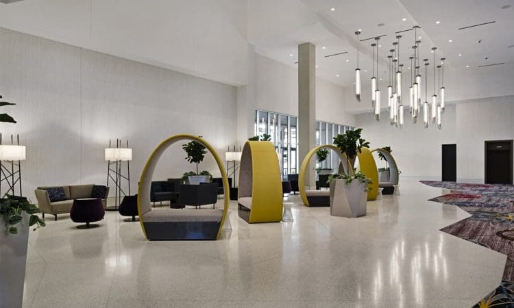 Modern hotel lobby with yellow circular seating pods, potted plants, lounge chairs, and contemporary hanging lights on a polished floor.