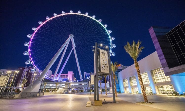 A large illuminated Ferris wheel is seen at night near modern buildings and palm trees, with signs reading "Caesars Forum" visible in the foreground.