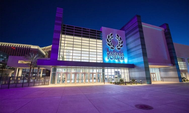 Exterior view of Caesars Forum convention center at night, illuminated with purple lighting, featuring large glass windows and the venue's logo above the entrance.