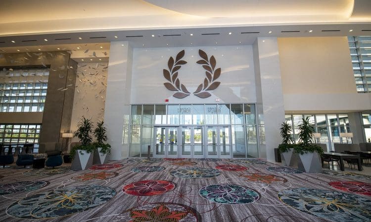 Spacious hotel lobby with patterned carpet, large indoor plants, floor-to-ceiling windows, and a wall decoration featuring a laurel wreath above glass doors.