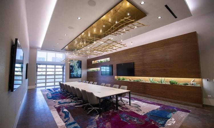 Modern conference room with a long white table, white chairs, large TV screens, wood accent wall, decorative lights, and a colorful abstract rug.