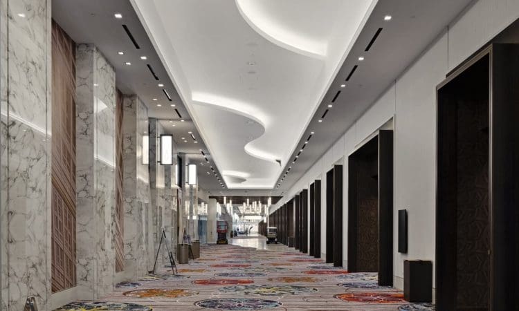 Modern hotel hallway with marble walls, a white curved ceiling, recessed lighting, and a colorful patterned carpet. Multiple doorways line both sides of the corridor.