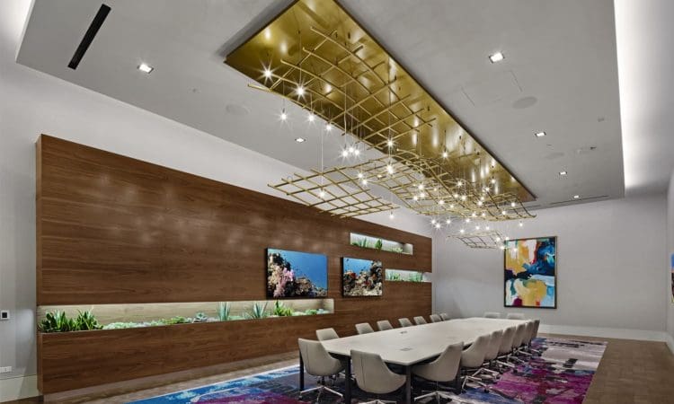 Modern conference room with a long white table, white chairs, wood-paneled wall with fish tanks, abstract art, colorful rug, and geometric gold ceiling light fixture.