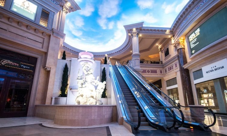 Interior construction work on Caesars Forum in Las Vegas with a sky-painted ceiling, a fountain statue, an escalator, and various storefronts including jeweler and fashion stores.