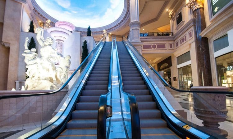 A pair of empty escalators in an ornate indoor mall with classical statues and columns under a painted sky ceiling.