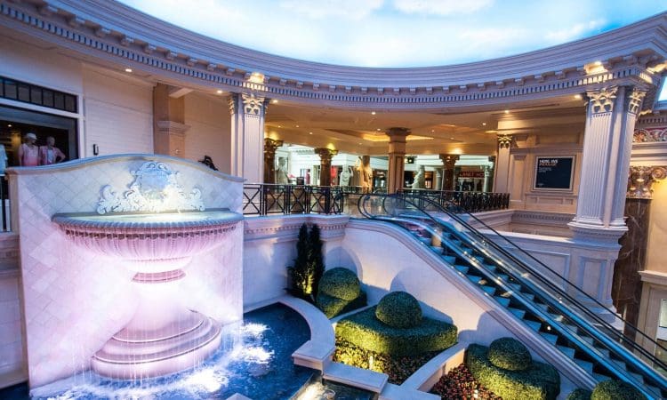 Luxurious indoor space featuring a large decorative fountain, manicured bushes, escalators, and classical architectural details with columns and ornate moldings.