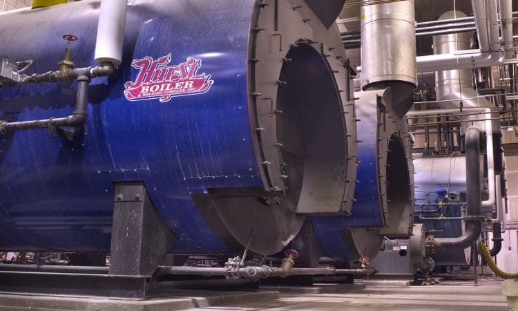 Large industrial boilers with pipes and valves are lined up in a facility. The nearest boiler has a red “Hurst Boiler” logo.