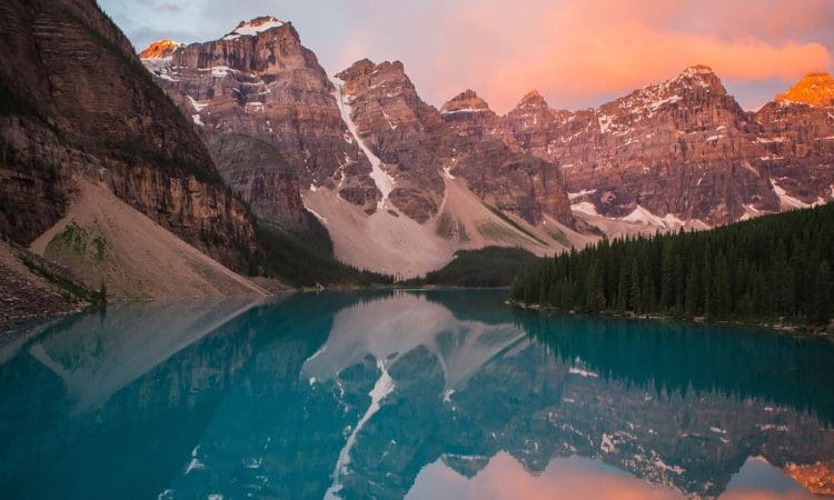 Mountain peaks reflected in a clear blue lake at sunrise, with patches of snow and dense pine trees along the shoreline.