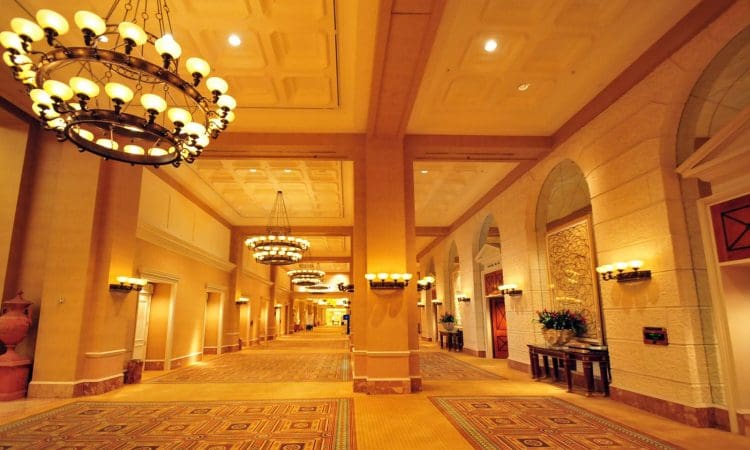 Spacious, well-lit hotel corridor with chandeliers, large columns, patterned carpet, and decorative wall features.