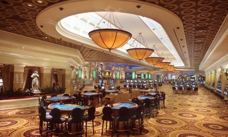 Casino interior with round tables, slot machines, ornate ceiling lights, decorative statues, and a patterned carpet. No people are present.