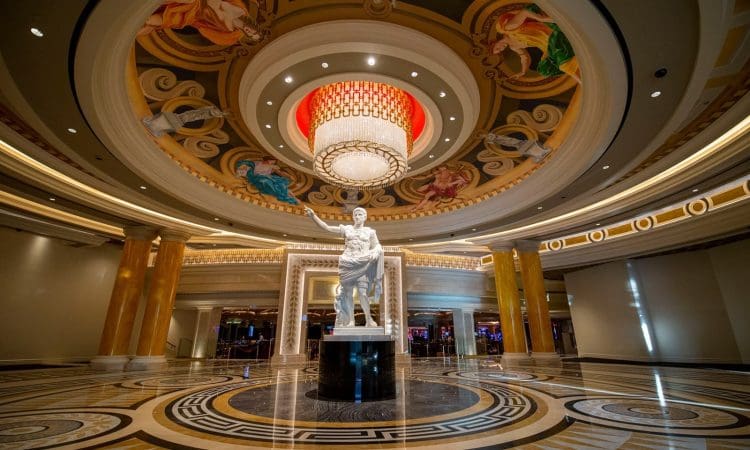 A grand rotunda features a white statue of a robed figure beneath a large chandelier and ornate ceiling with gold pillars and intricate artwork.